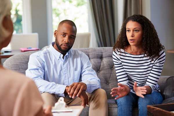 Pareja conversando con un terapeuta en un entorno acogedor, discutiendo temas emocionales y de relación. La mujer se expresa con gestos mientras el hombre escucha atentamente.