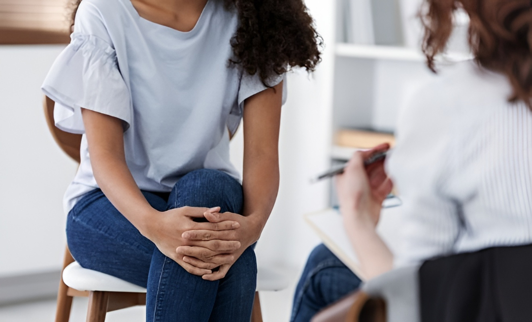 Mujer joven sentada en una silla durante una sesión de terapia, con una expresión reflexiva y las manos entrelazadas. En el fondo, se observa a otra persona tomando notas. La imagen transmite un ambiente de apoyo y comunicación en un contexto de salud mental.