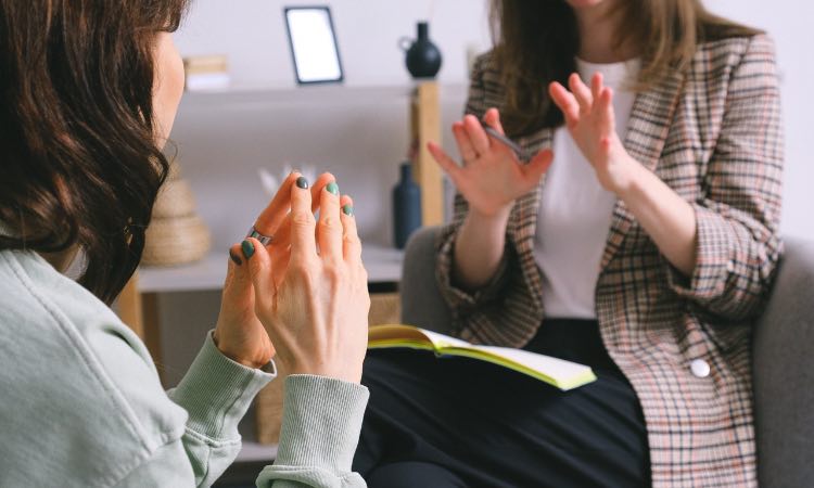 Dos mujeres en una conversación, una de ellas gesticulando con las manos mientras la otra escucha atentamente. Están sentadas en un ambiente moderno y acogedor, rodeadas de elementos decorativos. Una de las mujeres sostiene un cuaderno amarillo, sugiriendo una charla sobre desarrollo personal o terapia.